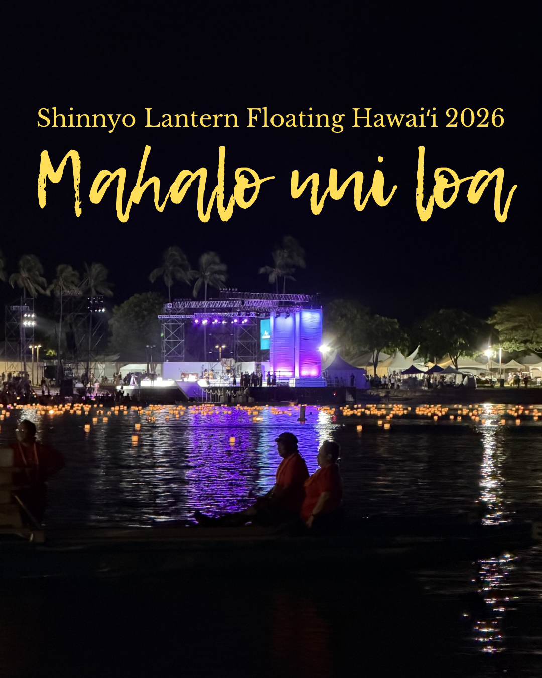 Nighttime image of the stage lit up in a soft purple light after the ceremony is over. It is across the water, and the water is filled with lit lanterns. In the front are the silhoutte of two figures on a canoe.